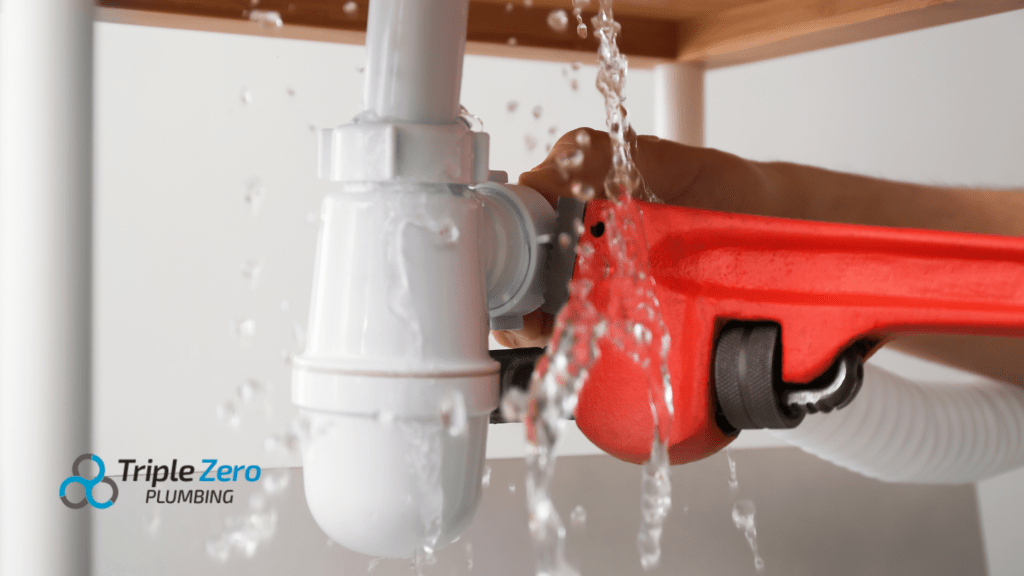 leaking white pipe being repaired with a red pipe wrench. Water is visibly spraying from the joint, indicating an active leak. The logo in the bottom-left corner reads "Triple Zero Plumbing."
